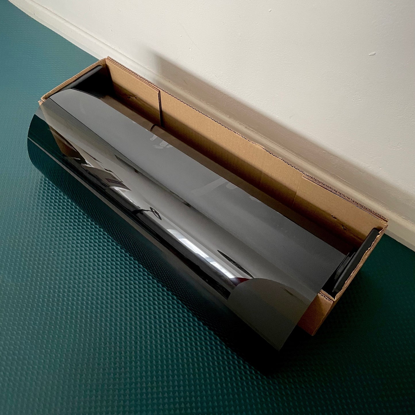 A roll of black automotive nano ceramic window film placed inside a cardboard box on a floor.